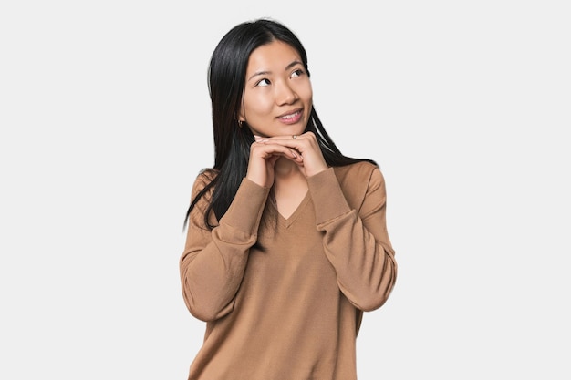 Young Chinese woman in studio setting keeps hands under chin is looking happily aside