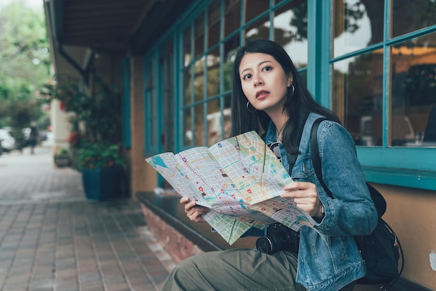 Young chinese woman tourist sitting outside restaurant holding\
paper map searching direction on bench chair after lunch in country\
style meal. independent female backpacker taking guide book\
visiting