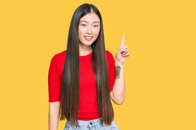 자신감 있고 행복한 미소를 지으며 캐주얼 옷을 입고 손가락 번호 1을 가리키는 젊은 중국 여성
