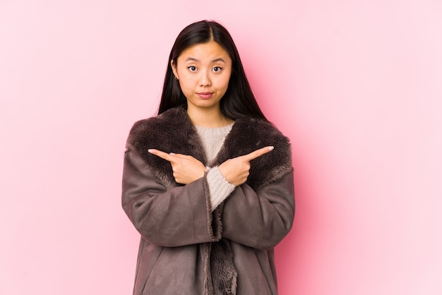 Young chinese woman wearing a coat isolated points sideways, is trying to choose between two options.