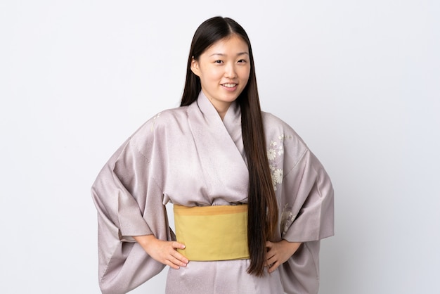Кимоно молодой китаянки над изолированной стеной позирует с руками на бедре и улыбается