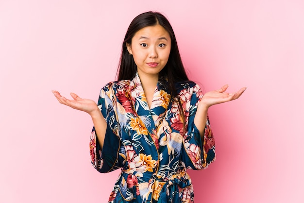 Young chinese woman wearing a kimono pajama isolated doubting and shrugging shoulders in questioning gesture.