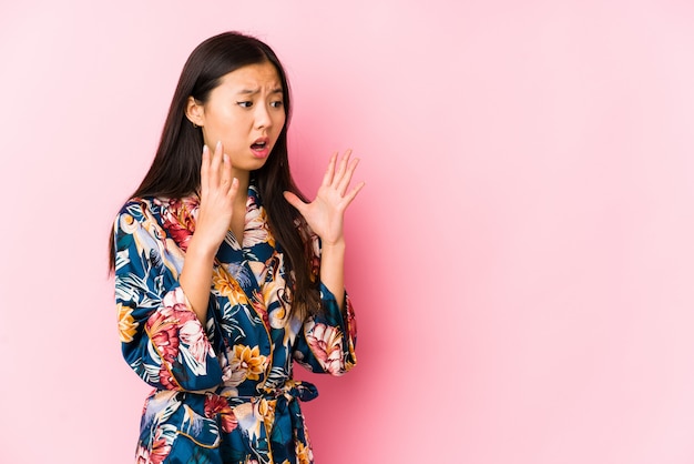 Young chinese woman wearing a kimono pajama isolated shouts loud, keeps eyes opened and hands tense.