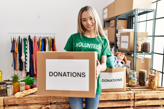 Young chinese woman wearing volunteer uniform holding donations box at charity center