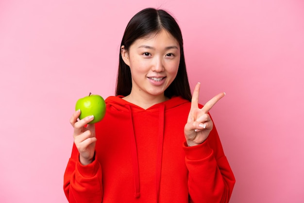 Young Chinese woman with an apple isolated on pink background smiling and showing victory sign