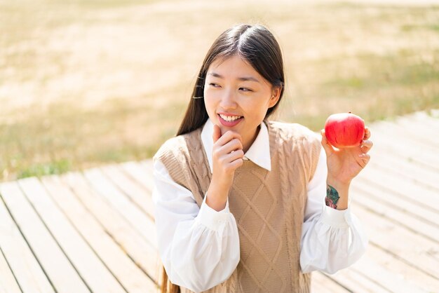 屋外でリンゴを持った若い中国人女性がアイデアを考え、側面を見ている