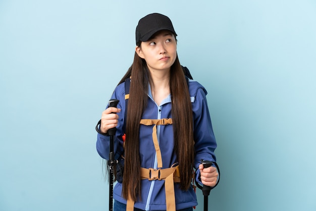 Young Chinese woman with backpack and trekking poles over isolated blue wall and looking up