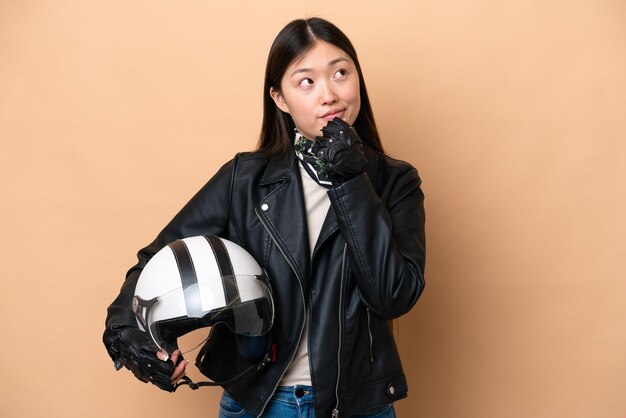 Young chinese woman with a motorcycle helmet isolated on beige\
background and looking up