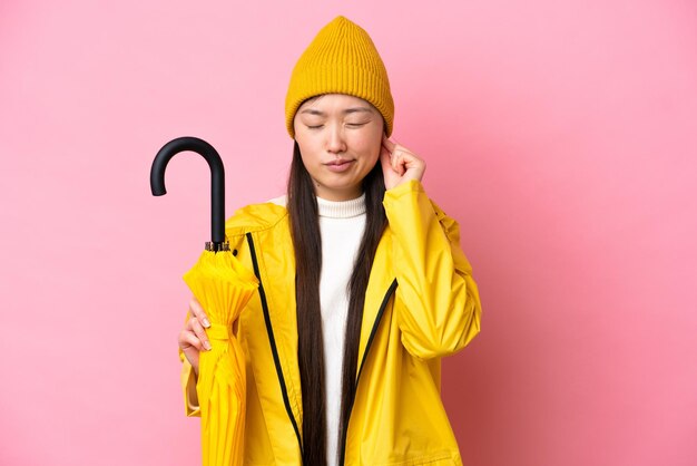 Young Chinese woman with rainproof coat and umbrella isolated on pink background frustrated and covering ears