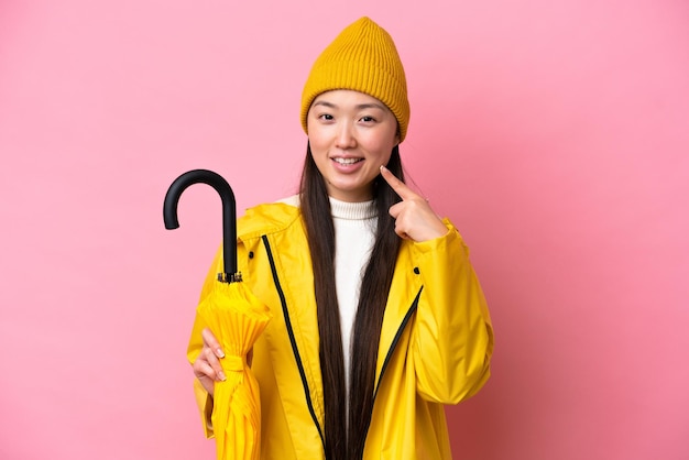 Young Chinese woman with rainproof coat and umbrella isolated on pink background giving a thumbs up gesture