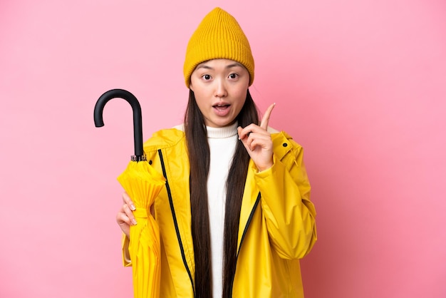 Young Chinese woman with rainproof coat and umbrella isolated on pink background intending to realizes the solution while lifting a finger up