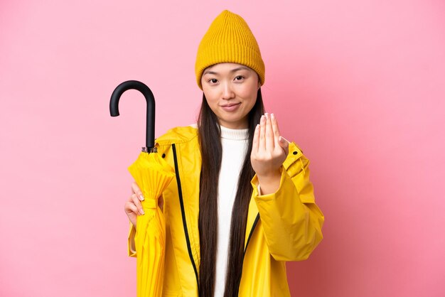 Young Chinese woman with rainproof coat and umbrella isolated on pink background inviting to come with hand Happy that you came