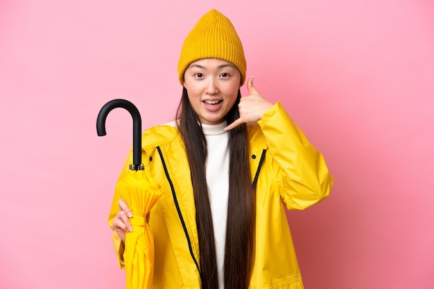 Young Chinese woman with rainproof coat and umbrella isolated on pink background making phone gesture Call me back sign