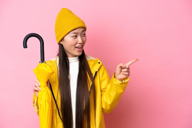 Young chinese woman with rainproof coat and umbrella isolated\
on pink background pointing finger to the side and presenting a\
product