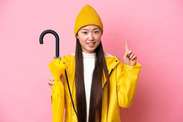 Young Chinese woman with rainproof coat and umbrella isolated on pink background pointing up a great idea