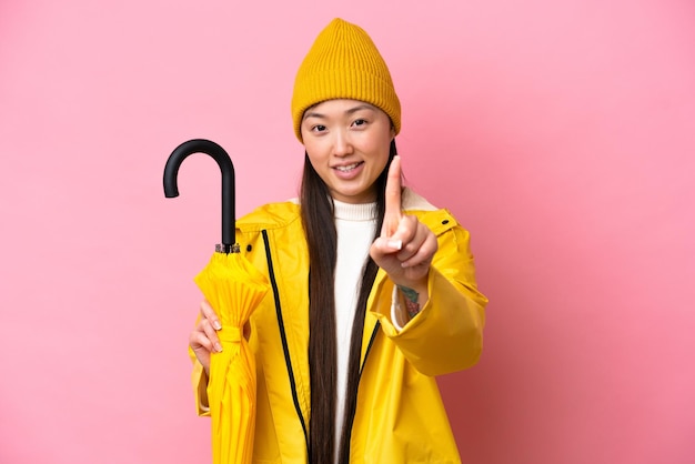 Молодая китаянка в непромокаемом пальто и зонтике на розовом фоне показывает и поднимает палец
