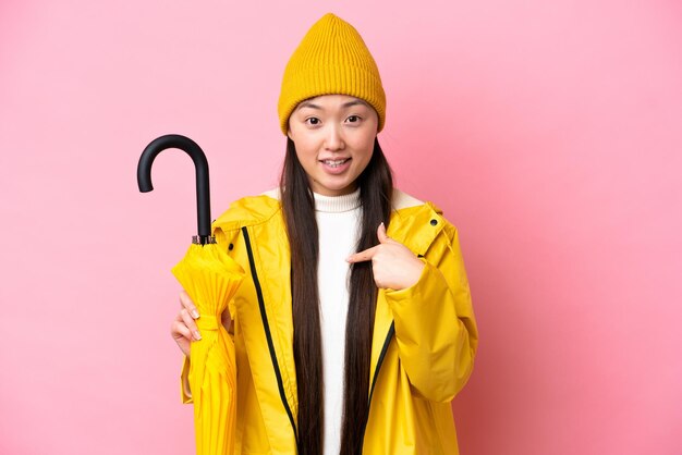 Young chinese woman with rainproof coat and umbrella isolated on pink background with surprise facial expression