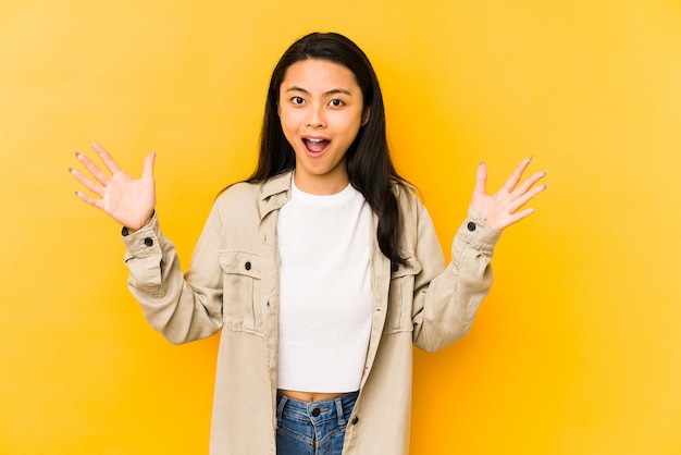 Young chinese woman on a yellow wall receiving a pleasant surprise, excited and raising hands.