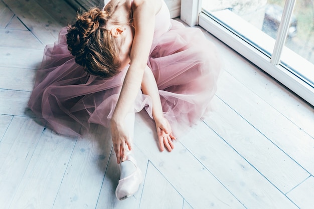 Young classical ballet dancer girl in dance class. Beautiful graceful ballerina in pink tutu skirt puts on pointe shoes