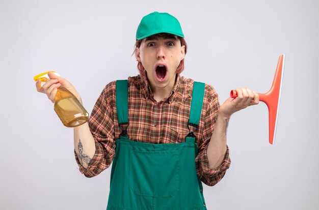 Young cleaning man in plaid shirt jumpsuit and cap holding cleaning spray and mop  amazed surprised standing over white wall