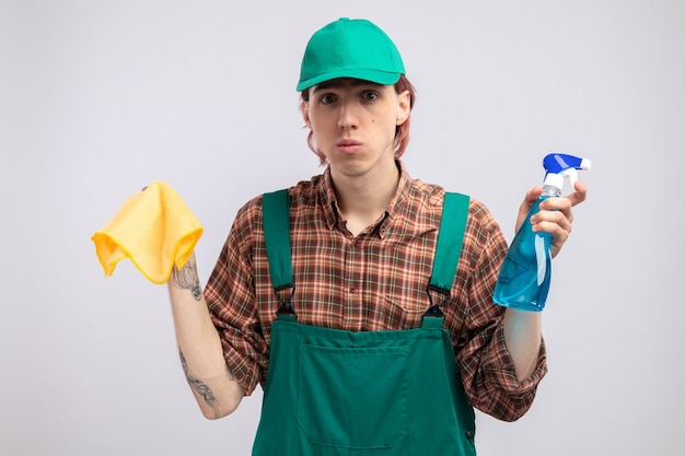 Молодой уборщик в клетчатой рубашке, комбинезоне и кепке, держит тряпку и чистящий спрей, смотрит с серьезным лицом