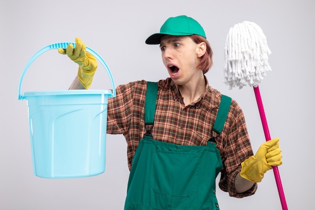 Young cleaning man in plaid shirt jumpsuit and cap wearing rubber gloves holding bucket and mop shouting being confused and surprised