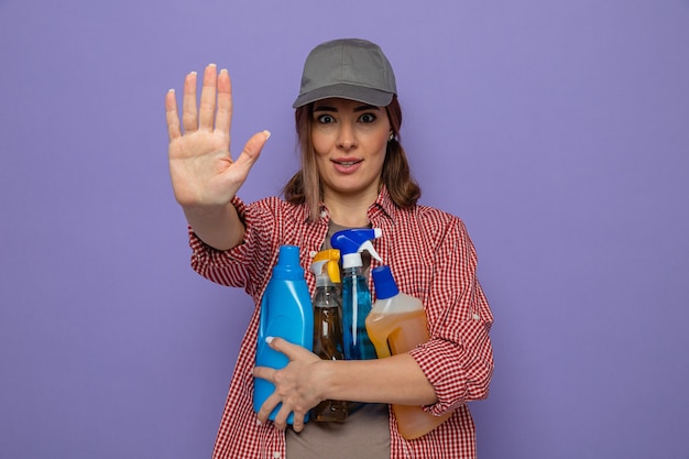Молодая уборщица в клетчатой рубашке и кепке, держащая бутылки с моющими средствами, смотрит в камеру с серьезным лицом, делая стоп-жест рукой, стоящей на фиолетовом фоне