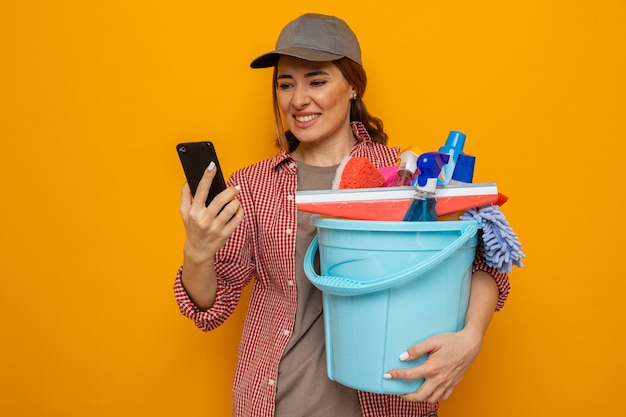 Giovane donna delle pulizie in camicia a quadri e cappello che tiene il secchio con strumenti per la pulizia guardando il suo telefono cellulare infastidito in piedi su sfondo arancione