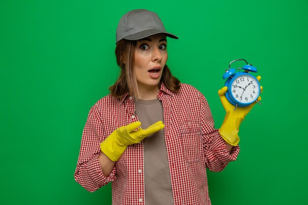 Foto giovane donna delle pulizie in camicia a quadri e berretto che indossa guanti di gomma tenendo la sveglia presentandola con il braccio guardando la telecamera confusa in piedi su sfondo verde