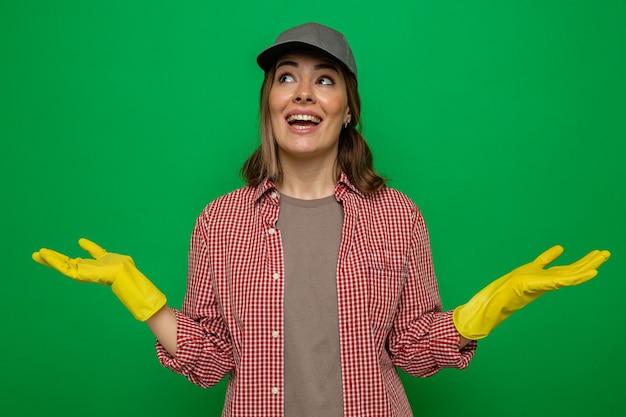 Giovane donna delle pulizie in camicia a quadri e berretto che indossa guanti di gomma che guardano sorridendo allegramente allargando le braccia ai lati