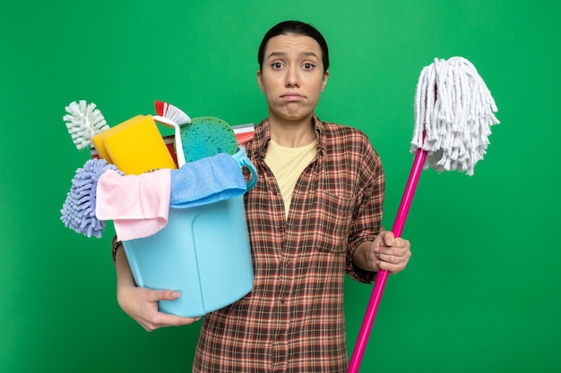 Young cleaning woman in plaid shirt holding bucket with cleaning tools and mop confused standing on green