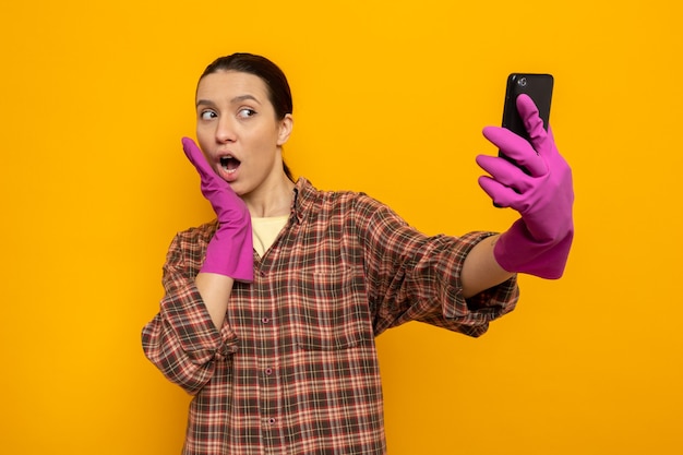 Young cleaning woman in plaid shirt in rubber gloves holding smartphone doing selfie happy and surprised standing on orange