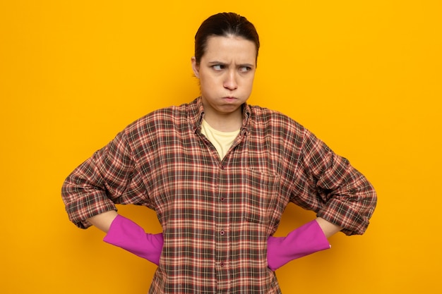 Young cleaning woman in plaid shirt in rubber gloves looking aside displeased blowing cheeks with arms at hip