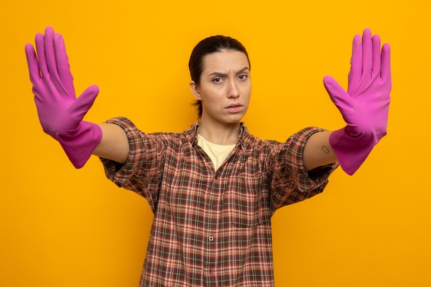 Young cleaning woman in plaid shirt in rubber gloves looking at front with serious face making stop gesture with hands standing over orange wall