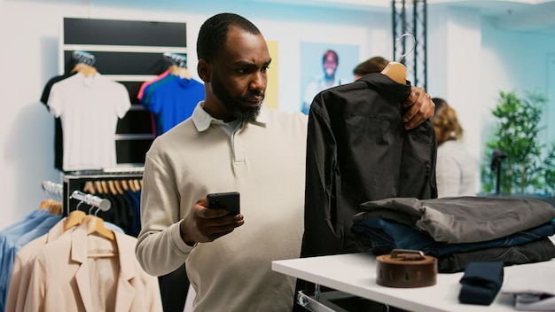 Young client analyzing online website app to find clothes, buying new fashion collection in department store. Customer with mobile phone checking merchandise of formal wear, small business.