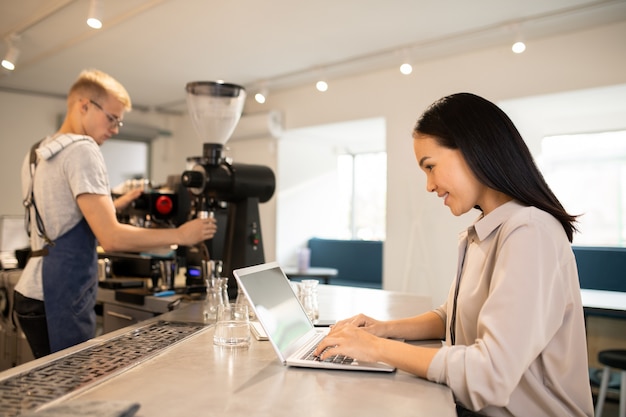 Young client of cafeteria sitting by table and surfing in the net while barista preparing fresh coffee on background