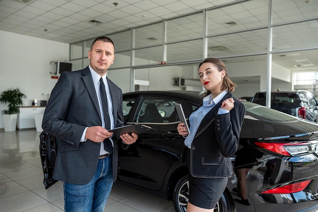 クリップボードを保持しているサロンのセールスマンで新車の近くに自動車ディーラーを持つ若いクライアント