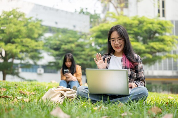 젊은 대학생이 공원에서 앉아있는 동안 노트북으로 비디오 통화 회의를 하고 있습니다.