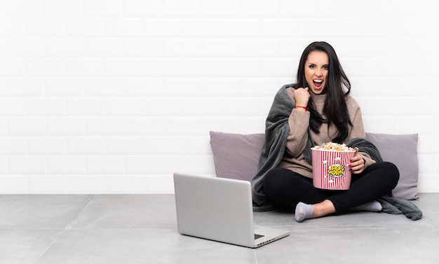 Young Colombian girl holding a bowl of popcorns and showing a film in a laptop frustrated by a bad situation
