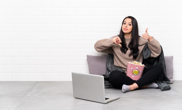 Young Colombian girl holding a bowl of popcorns and showing a film in a laptop making good-bad sign. Undecided between yes or not