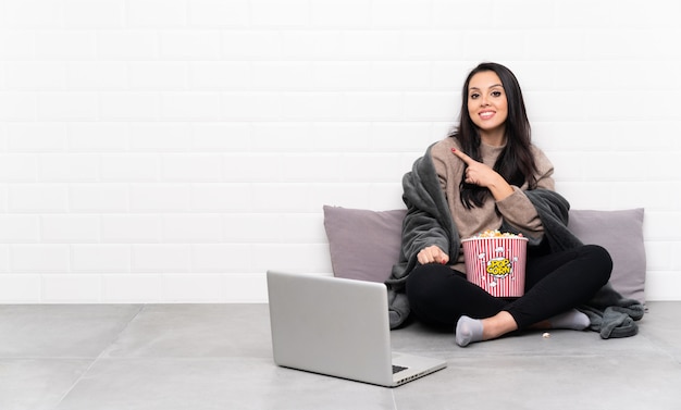 Young Colombian girl holding a bowl of popcorns and showing a film in a laptop pointing to the side to present a product