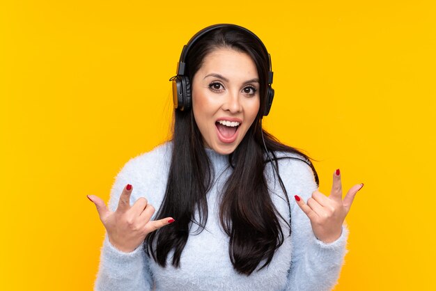 ロックジェスチャーを作る音楽を聞いて孤立した黄色の壁の上のコロンビアの少女