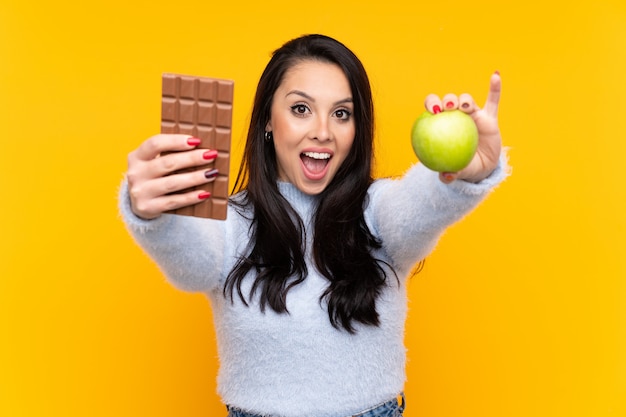 Young Colombian girl over isolated yellow wall taking a chocolate tablet in one hand and an apple in the other