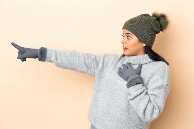 Photo young colombian girl with winter hat on beige wall pointing finger to the side