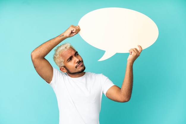 Young Colombian handsome man isolated on blue background holding an empty speech bubble and with sad expression