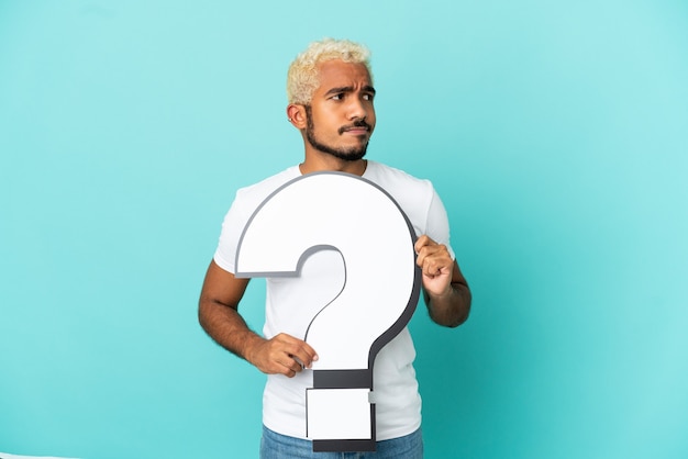 Photo young colombian handsome man isolated on blue background holding a question mark icon and looking side