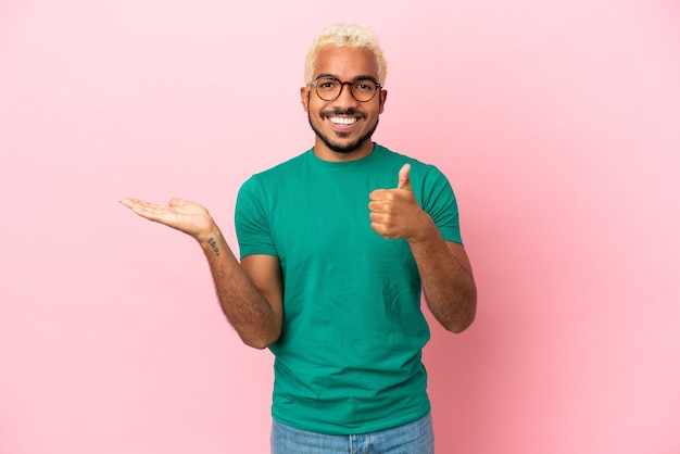 Young Colombian handsome man isolated on pink background holding copyspace imaginary on the palm to insert an ad and with thumbs up