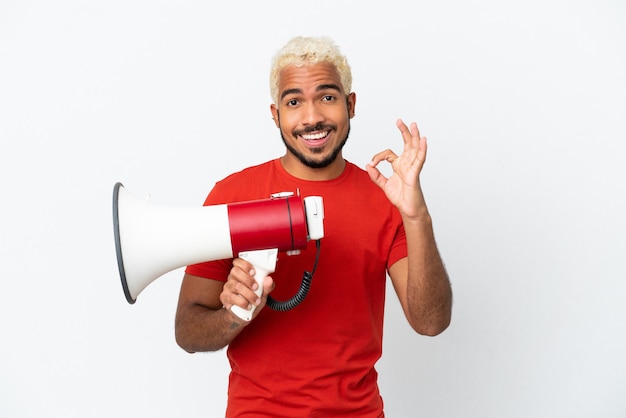Young Colombian handsome man isolated on white background holding a megaphone and showing ok sign with fingers