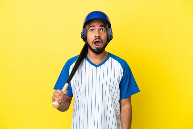 Young Colombian latin man playing baseball isolated on yellow background looking up and with surprised expression