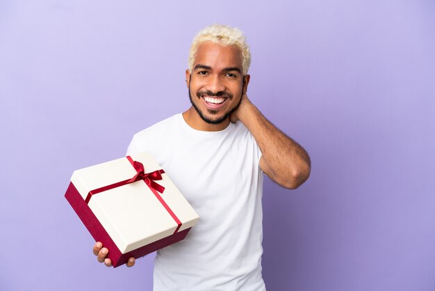 Young Colombian man holding a gift isolated on purple background laughing